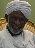 This photograph features the late Hassan al-Turabi with a warm smile and a neatly groomed beard, wearing a white turban and traditional Sudanese attire. He is also wearing glasses, which frame his cheerful and kind expression. The background shows part of a golden, ornate chair with striped upholstery, adding a touch of elegance to the setting.