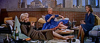 Monroe en How to Marry a Millionaire. Lleva un traje de baño naranja y está sentada junto a Betty Grable, que lleva pantalones cortos y una camisa, y Lauren Bacall, que lleva un vestido azul.
