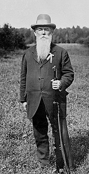 Photographie montrant Oscar Swahn debout, portant une veste, un chapeau, une barbe et une moustache et tenant une carabine dans la main gauche.