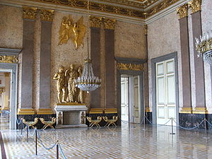 Farbfotografie eines Innenraums mit drei goldenen Figuren auf dem Kamin der hinteren Wand. Über ihnen befinden sich zwei goldene Engel, die an beiden Seiten jeweils von zwei grauen Säulen mit goldenen Enden gesäumt werden. Unter den Säulen stehen vier Hocker und an den Wandseiten sind zwei offene Türen mit goldenen Ornamenten. An der rechten Wand befinden sich eine verschlossene Tür und zwei Säulen. Der gold-weiße Boden ist an beiden Seiten von Absperrkordeln begrenzt.