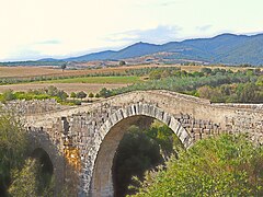 Photographie d'un ancien pont à arches avec un paysage agricole et champêtre ainsi que des collines en arrière-plan.