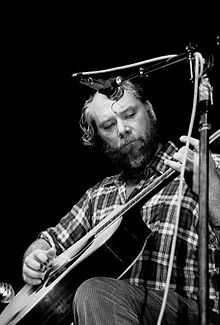 Positioned behind two microphones, a bearded man dressed in a flannel shirt and corduroy pants fingerpicks an acoustic guitar.