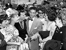 Black-and-white photograph of a young man wearing a suit and bow tie standing with several smiling ladies, with a crowd of more fans, journalists, and police officers in the background