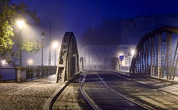 Vista noturna da ponte Młyński norte (36,6 metros), sobre o braço norte do rio Óder em Breslávia, Polônia. A ponte conecta a margem direita do rio com a ilha Młyńska no centro histórico da cidade. Existem dois trilhos de bonde através da ponte, tráfego de carros nos dois sentidos (a carga máxima da ponte é definida em 10 toneladas) e pedestres (as calçadas são separadas da estrada e da linha férrea com uma armação de suporte de carga). Os danos à ponte durante o cerco à cidade no final da Segunda Guerra Mundial foram reparados temporariamente com vigas de madeira logo após a guerra. A renovação subsequente em 1956, durante a qual a plataforma de aço foi reconstruída e as vigas reforçadas, deu-lhe um traçado viário moderno. (definição 4 548 × 2 806)