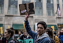 Demonstration. Im Vordergrund ein junger schwarzer Mann mit offenen Mund, der mit nach oben ausgestreckten Arm ein Pappschild in die Bildmitte hält mit der Aufschrift: „FUCK RACIAL PROFILING“. Auf dem Pappschild ist ein schwarze Gesichtsfläche rechts und links das gleiche Schattenbild gespiegelt. Allerdings ist dort der Hintergrund schwarz und die Fläche des Kopfes farblich ausgespart.