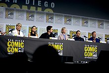 Linda Hamilton, Natalia Reyes, Diego Boneta, Mackenzie Davis, Gabriel Luna and Arnold Schwarzenegger seated at a table on a dais.