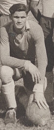 Black-and-white photo of Max Wosniak on one knee in the grass. One of his hands is resting on a soccer ball on the ground.