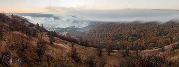 Le creux d'une vallée arrondie, composé d'arbustes et d'herbes jaunies, donnant sur une forêt en automne. Plus loin les nuages enveloppent les collines, laissant la place au ciel, d'où pointe une douce lumière orangée.