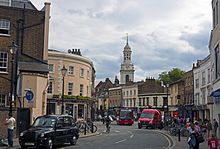 A curving street with older two- and three-storey buildings on either side. In front is a black London taxicab with an advert; midway down the street is an intersection with heavy traffic. A cupolaed clock tower rises in the rear