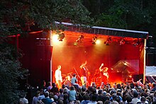 Foto einer Konzertbühne, die rot ausgeleuchtet ist. Auf der Bühne stehen winkende Musiker, davor sieht man dicht gedrängt die Köpfe von Zuschauern