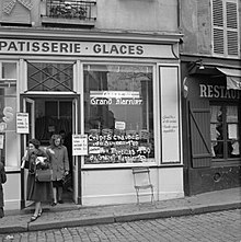 Boulangerie typique parisienne en 1965, d'où laquelle sortent deux clientes. Dans la vitrine, une enseigne lumineuse porte l'inscription crêpes au Grand Marnier, informant les passants que ces produits sont proposés à la vente en ce lieu. D'autres inscriptions en indiquent aussi le prix à l'époque : un franc cinquante.