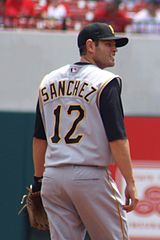 A man wearing a gray baseball uniform with the name "Sanchez" and the number "12" on the back, a black cap bearing, and a baseball glove stands on a baseball field