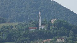 Six buildings peek out of lush green trees, with a hill behind. A tall belltower stands in the middle.