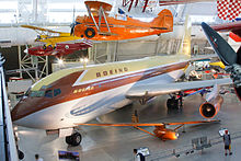 Vue plongeante, de trois-quarts avant, d'un avion de couleur jaune, marron et gris dans le hall d'un musée ; plusieurs autres avion, plus petits, sont exposés tout autour, au sol ou suspendus en l'air.