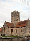 Brown stone building with red roofs and central square tower. In the foreground are gravestones.