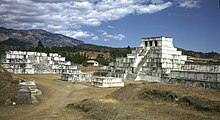 Un grupo de pirámides escalonadas blancas, el más alto de ellos coronado por un santuario con tres entradas. En el fondo, una cresta de montañas bajas.