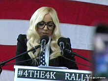 Uma mulher com cabelo loiro falando em um pódio em vários microfones. Ela usa óculos grandes. O fundo é uma série de listras horizontais vermelhas e brancas.