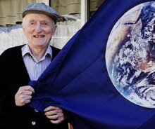 Elderly man in a collared shirt and hat standing, holding a blue flag with a picture of planet Earth on it