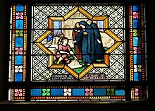 Photographie en couleur d'un vitrail figurant un groupe de personnes autour d'un plan, avec une église en arrière-plan.
