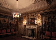 A photograph of a richly-decorated room, with a white marble fireplace surrounded by elaborate wall carvings of twisted pillars and scrolling foliage. The ceiling is also decorated with plaster fruit and flowers. Tapestries line the walls, and a set of satin-covered chairs are placed at the edge of the room.