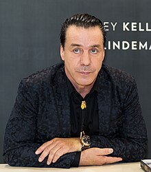 Lindemann trägt dunkle Kleidung und ist sitzend abgebildet. Das dunkle Haar ist kurz.