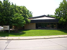 A one-story sandstone building with a gently sloping roof sits behind a green lawn and some dense trees.