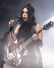 Chelsea Wolfe, Sängerin, mit Gitarre im schwarzen Kleid vor einem Mikrofon auf der Bühne. Sie hat schwarze, kinnlange Haare und sing gerade.