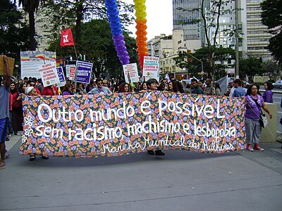 Grupo de mulheres marchando pela calçada segurando uma faixa com os dizeres “Outro mundo é possível sem racismo, machismo e lesbofobia - Marcha Mundial das Mulheres”