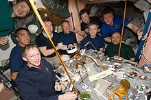 Nueve astronautas alrededor de una mesa con varias latas de comida. En el fondo, se ve una selección de equipos y las paredes color salmón del nodo Unity.