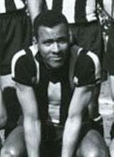 A young man sitting down, wearing a striped shirt. Behind him, three men wearing the same shirt and dark shorts are partially visible