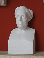 Buste (statue) de Noether en plâtre posé sur une dalle en marbre devant un mur rouge.