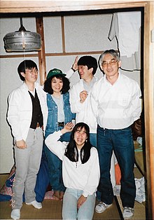 Três homens orientais e uma mulher oriental em pé, uma mulher oriental ajoelhada, todos sorrindo em pose de fotografia, numa sala de estar em osaka, Japão