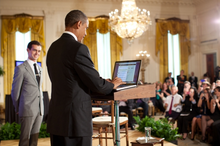 Man in his twenties smiling at left, man in his forties using computer at center, large crystal chandelier, several people in audience