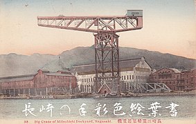 Carte postale montrant une gigantesque grue en métal. Elle domine de très grands hangars industriels, des montagnes sont visibles au loin.