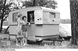 Photo noir et blanc de 1958 ; trois femmes discutent sur le pas d'une petite caravane blanche installée sous des arbres, non loin d'un lac.