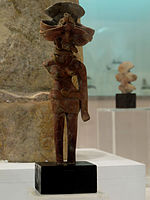تمثال الإلهة الأم يرتدي سترة قصيرة ضيقة مع تنورة مثبتة بحزام على الخصر باستخدام ميدالية مثل المشبك وسروال ضيق وقلائد وزخارف للأذن.