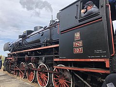 Zdjęcie przedstawia lokomotywę parową stojącą na stacji w Żabnie. Lokomotywa pomalowana jest na czarno i czerwono, a z pod jej kół i z gwizdka wylatuje para. W oknie lokomotywy widać maszynistę ubranego w niebieskoszarą koszule.