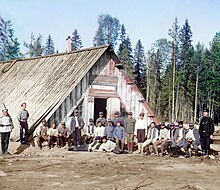Barevná fotografie skupiny rakousko-uherských zajatců seřazených před dřevěným stavením. Po krajích je hlídá trojice ruských vojáků.