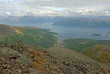 Foto von einem Berg herunter auf eine Ortschaft, die an einem Fjord liegt. Auf der anderen Fjordseite ebenfalls hohe Erhebungen