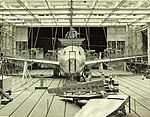 Cliché noir et blanc. Hangar avec de très nombreux éléments d'assemblage. Au centre avion en cours de montage. Pas de nez.