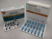 Η χαρτονένια συσκευασία των δύο φαρμάκων που χρησιμοποιούνται για τη θεραπεία της παχυσαρκίας. Η ορλιστάτη φαίνεται πάνω με εμπορική επωνυμία Xenical σε λευκή συσκευασία με το λογότυπο της Roche στην κάτω δεξιά γωνία (η επωνυμία της Roche μέσα στο εξάγωνο). Η σιβουτραμίνη είναι από κάτω με την εμπορική επωνυμία Meridia. Η συσκευασία είναι λευκή στην κορυφή και μπλε στο κάτω μέρος και διαχωρίζεται με μετρητική ταινία. Το Α του λογοτύπου των Εργαστηρίων Abbott βρίσκεται στο κάτω μισό της συσκευασίας.