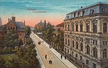 Carte postale ancienne colorisée montrant une large allée bordée d'arbres et un grand bâtiment sur la droite.