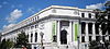 The corner of a white stone, classically designed, 4 story building. The door is on the closer side of the corner, is on the second story with a grand staircase up to it, and is flanked by two ionic columns.