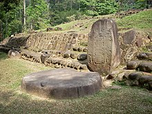 A squat standing stone with a flat front face set against a low stone-faced mound structure at right. The face of standing stone is inscribed but the details are not visible. In front of the stone, at left, lies a flat circular stone, set upon a flat grassy area. Dense vegetation is visible in the background.