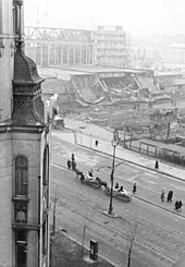 Czarno-białe zdjęcie przedstawiające widok fragmentu Alej Jerozolimskich i, na dalekim planie, ruin dworca. Na pierwszym planie po lewej, wzdłuż boku zdjęcia, widoczny wykusz jednego z domów przy Al. Jerozolimskich. Dolną część zdjęcia zajmuje widok fragmentu ulicy, którą posuwa się grupa pieszych i dwa wozy konne wyładowane workami. Na dalekim planie widoczna bryła dworca, z częściowo odsłoniętymi elementami stalowej konstrukcji i zawalonymi elementami w przyziemiu. Najdalszy plan zamglony.