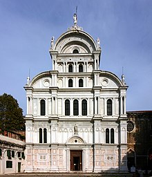 Image d'une façade d'église montrant un stylobate avec des éléments gothiques surmonté d'une série de petites niches aveugles et d'une arche au-dessus de laquelle s'appuient les contreforts latéraux sculptés. Dans les sections centrale et latérale, séparées par des doubles colonnes, se trouvent les fenêtres en séquence alternée, qui s'élèvent jusqu'à la rosace centrale. Dans la partie inférieure, de part et d'autre du portail surmonté d'une statue, quatre prophètes en buste sont insérés dans des cadres décorés de putti.