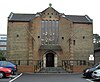 Front view of a brown- and red-brick building with five steps up to a round-arched window, above which are five narrow, tall rectangular windows. A shallow tiled roof rises behind. The steps are flanked by black railings and low brick walls. Cars are parked on both sides.