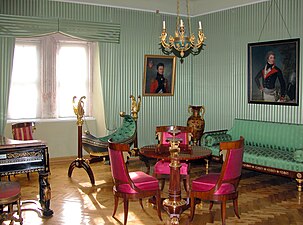 Farbige Eckansicht eines Innenraums mit grüngestreiften Wänden und Fenstervorhängen. Rechts vom Fenster befinden sich zwei Porträts von Militärangehörigen. An der rechten Wand stehen ein grünes Sofa und eine dunkle Vase. In der Mitte sind ein runder Tisch mit einer Etagere und drei Stühlen sowie eine grün gepolsterte Wiege aus Holz, die von goldenen Schwänen gehalten wird. Links ragt der Teil eines Klaviers in das Bild hinein.