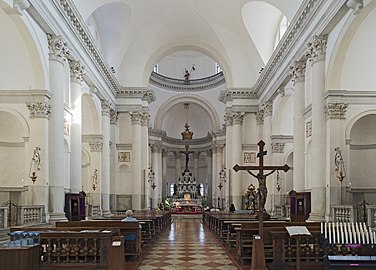 Image de l'intérieur d'une nef d'église avec des colonnades et arches de chaque côté, un pavement à damier, des bancs de chaque côyé de l'allée centrale, un grand crucifix au premier plan sur le côté gauche, une coupole en hauteur et au fond un autel.