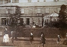 Photographie ancienne sur laquelle plusieurs enfants joue dans le jardin à l'arrière d'un manoir avec baie vitrée et véranda imposante.